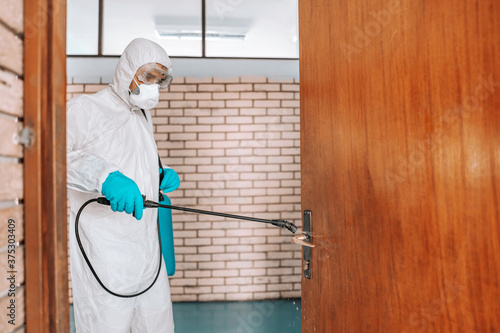 Worker in white sterile uniform, with rubber gloves and mask on holding sprayer with disinfectant and sterilizing doors in school.