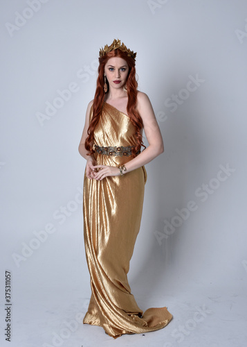 Full length portrait of girl with red hair wearing long grecian toga and golden wreath. Standing pose iisolated against a grey studio background.