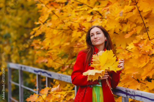 Portrait of pretty young woman of Slavic appearance in yellow colored dress and red coat in autumn, walk in forest. Cute model walks in Park in golden autumn against background of nature