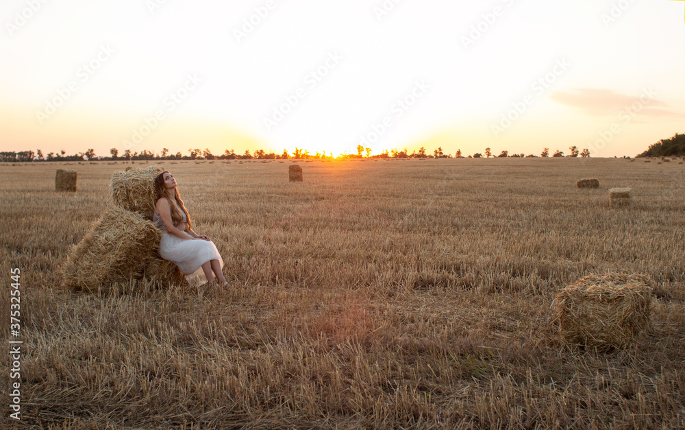woman sitting on hay stack walking in summer evening, beautiful romantic girl with long hair outdoors in field at sunset