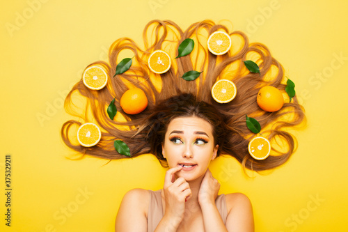 confused girl lying on studio background with orange fruits on long hair  young woman head thinking  making choice  view from above