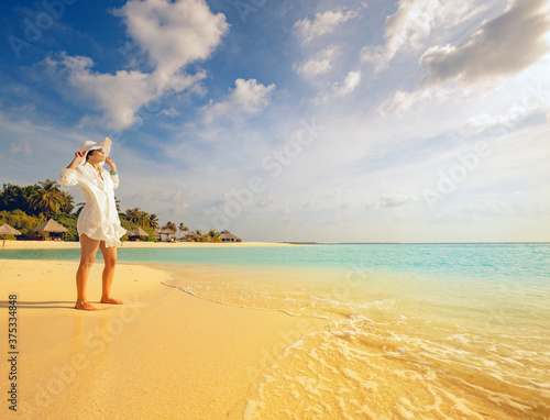 stylish girl watches the sunset on a tropical palm fringed beach of a maldives island with white hat and shirt with feet in soft sand