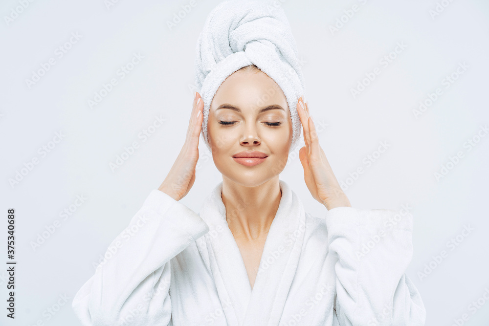 Gorgeous spa woman stands with closed eyes, keeps hands on towel wrapped on head, dressed in white bath robe, has healthy skin, natural makeup, well groomed complexion, poses indoor. Beauty treatment Gorgeous spa woman stands with closed eyes, keeps hands on towel wrapped on head, dressed in white bath robe, has healthy skin, natural makeup, well groomed complexion, poses indoor. Beauty treatment