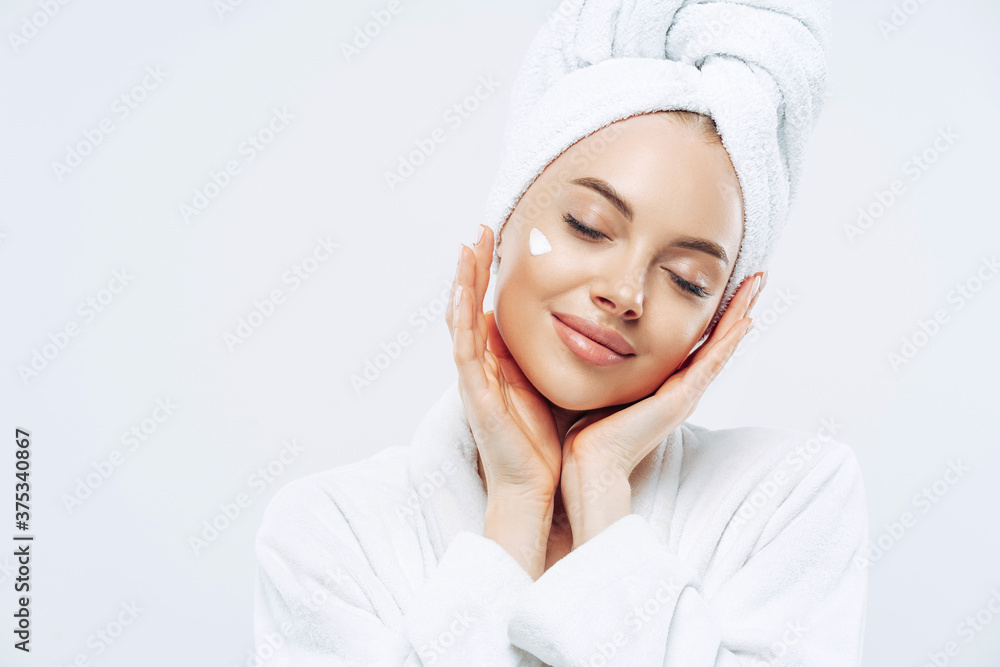 Studio shot of relaxed beautiful woman with closed eyes tilts head, touches skin gently, applies face cream, wears bath towel on head after taking spa procedures, enjoys facial treatments, body care Studio shot of relaxed beautiful woman with closed eyes tilts head, touches skin gently, applies face cream, wears bath towel on head after taking spa procedures, enjoys facial treatments, body care