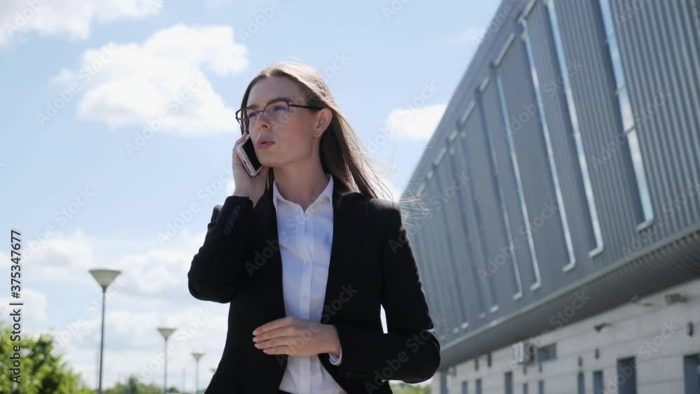 Beautiful and stylish business woman goes to work on the background of the large glass building.She speaks on the phone using wireless headphones. Beautiful and stylish business woman goes to work on the background of the large glass building.She speaks on the phone using wireless headphones.