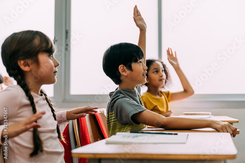 Elementary age Asian student boy raised hands up in Q and A class. Diverse group of pre-school pupils in elementary age in education building school. Volunteering and participating classroom concept.