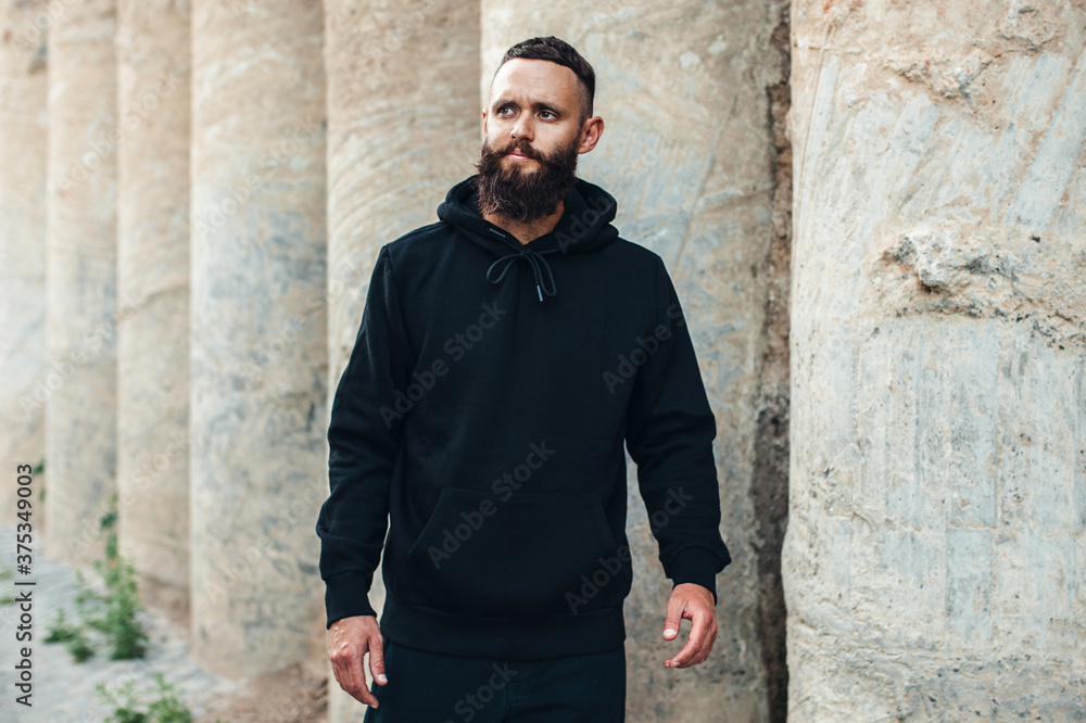 City portrait of handsome hipster guy with beard wearing black blank hoodie or sweatshirt with space for your logo or design. Mockup for print