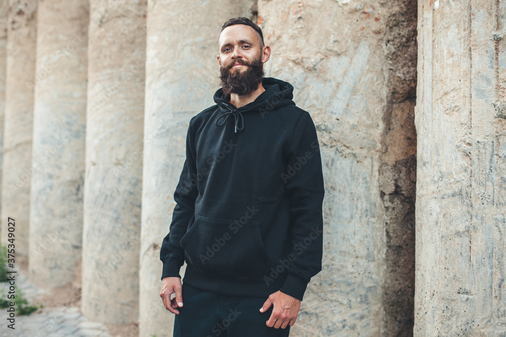 City portrait of handsome hipster guy with beard wearing black blank hoodie or sweatshirt with space for your logo or design. Mockup for print