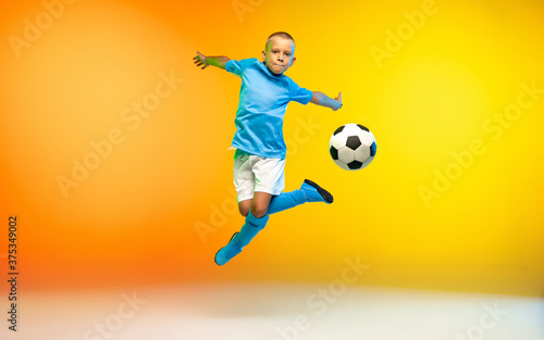 In jump. Young boy as a soccer or football player in sportwear practicing on gradient yellow studio background in neon light. Fit playing boy in action, movement, motion at game. Copyspace. © master1305