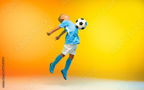 In jump. Young boy as a soccer or football player in sportwear practicing on gradient yellow studio background in neon light. Fit playing boy in action, movement, motion at game. Copyspace.
