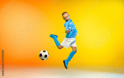 Dynamic. Young boy as a soccer or football player in sportwear practicing on gradient yellow studio background in neon light. Fit playing boy in action, movement, motion at game. Copyspace. © master1305