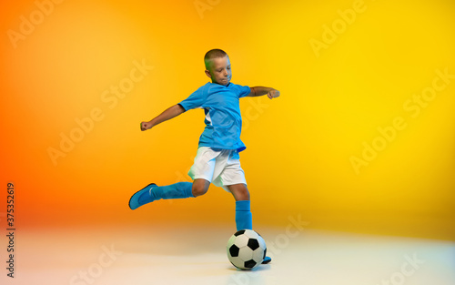 Kicking. Young boy as a soccer or football player in sportwear practicing on gradient yellow studio background in neon light. Fit playing boy in action, movement, motion at game. Copyspace. © master1305