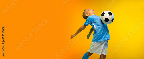 Winner. Young boy as a soccer or football player in sportwear practicing on gradient yellow studio background in neon light. Fit playing boy in action, movement, motion at game. Flyer, copyspace.