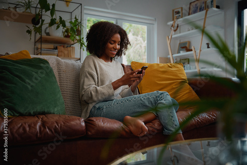 Happy young african woman using mobile phone while sitting a couch at home