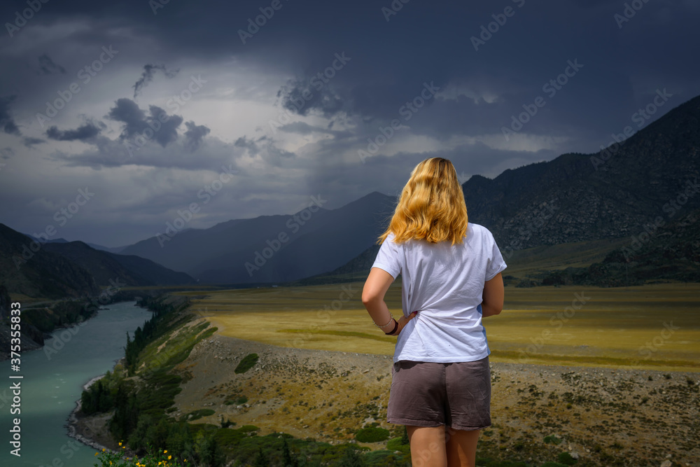 Young woman in white t-shirt standing back to the camera in the sunlight and looks at the beautiful view of Katun river, plains and rocks. In the distance, torrents of rain, overcast sky.