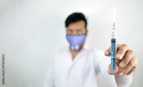 a medical professional in white coat and protective mask holding syringe in hand with selective focus in hand with blurred background