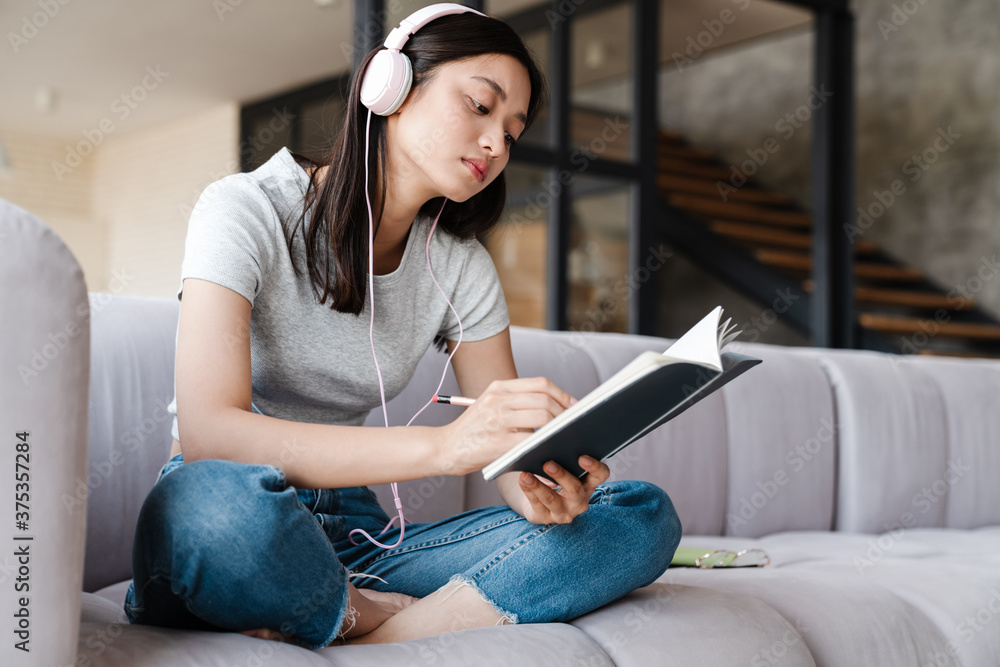 Obraz premium Image of focused asian woman using headphones and writing down notes