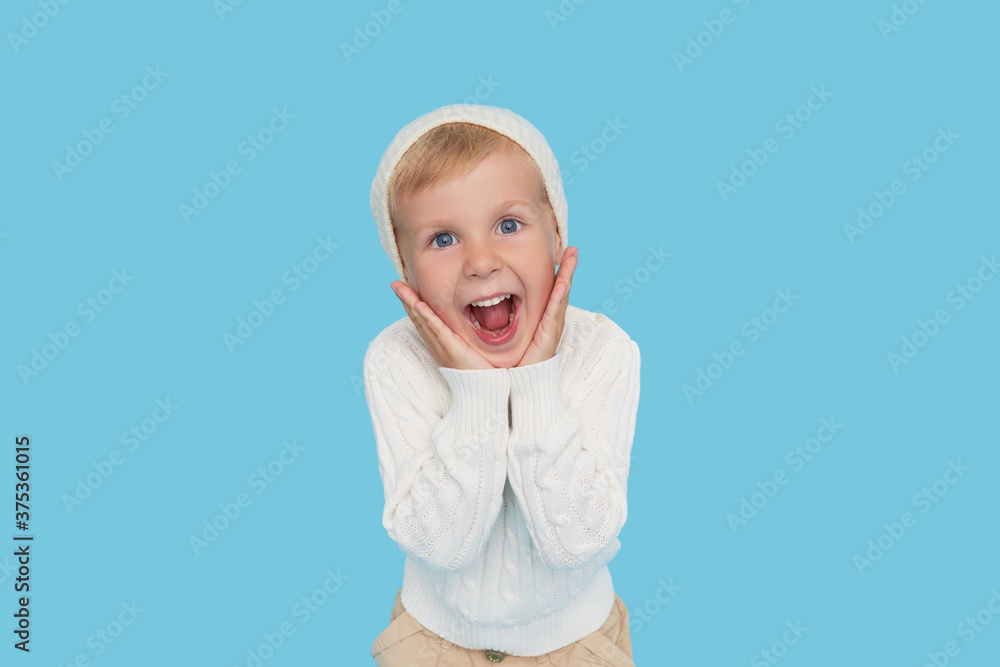 Wow face. Cute little blond boy in a warm white hat and sweater on a colored blue background. Winter season of childrens sales and discounts Wow face. Cute little blond boy in a warm white hat and sweater on a colored blue background. Winter season of childrens sales and discounts
