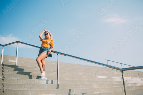 Trendy dressed amateur photographer with old fashioned technology camera standing at urban stairs, youthful hipster girl 20 years old looking away during recreation leisure at street setting