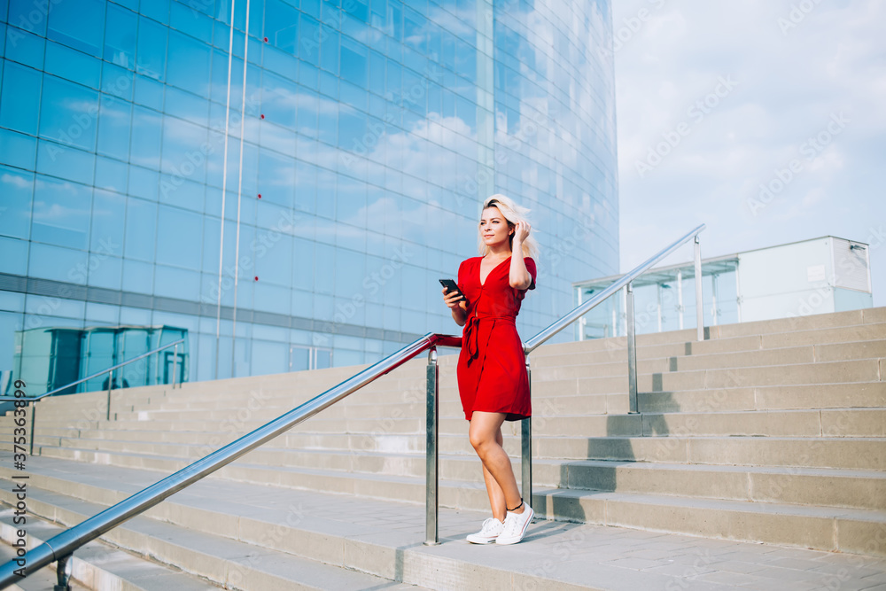 Young woman in stylish red sundress standing at urban stairs pondering on content idea for network sharing, millennial hipster girl holding modern cellular gadget in hand spending leisure for mobility