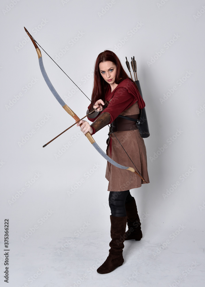 Full length portrait of girl with red hair wearing brown medieval archer costume.. Standing pose holding a bow and arrow, isolated against a grey studio background. Full length portrait of girl with red hair wearing brown medieval archer costume.. Standing pose holding a bow and arrow, isolated against a grey studio background.