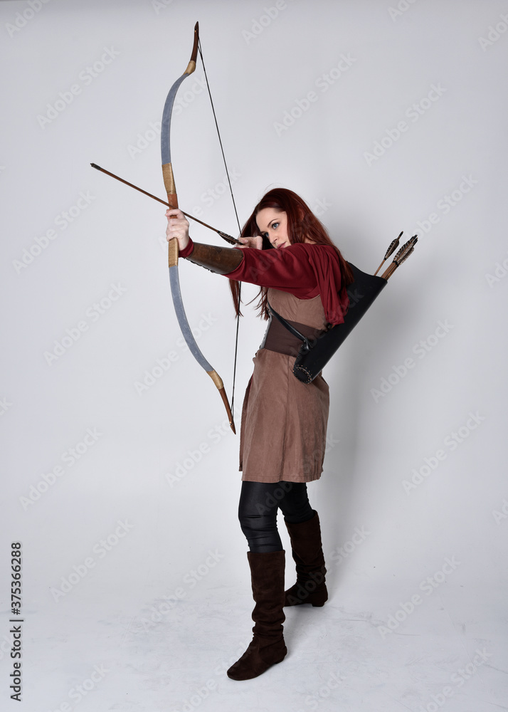 Full length portrait of girl with red hair wearing brown medieval archer costume.. Standing pose holding a bow and arrow, isolated against a grey studio background. Full length portrait of girl with red hair wearing brown medieval archer costume.. Standing pose holding a bow and arrow, isolated against a grey studio background.