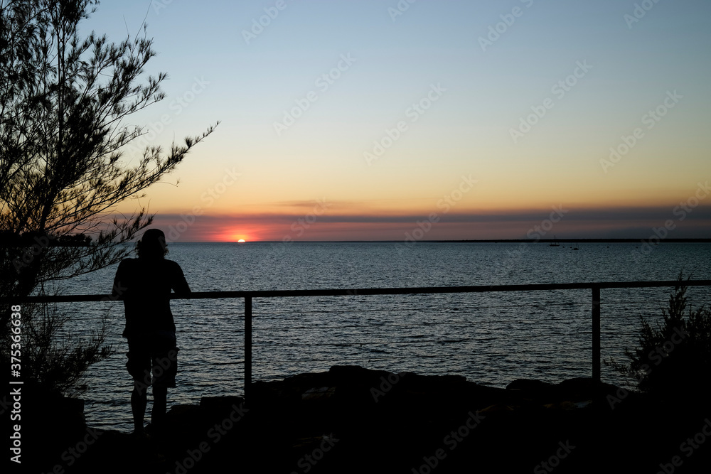 Silhouette of man leaning on a fence to watch the sunset over a large body of water.