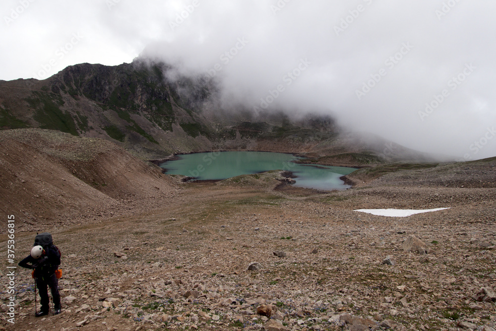 Alpine lake in the clouds. A tourist climbs the pass against the background of a high-mountain lake. Alpine lake Giybashkel (3240 meters above sea level), Caucasus. 