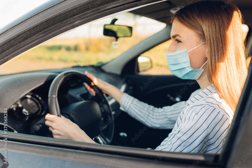 Beautiful young woman in a protective medical mask on her face, driving a car during a trip, travel concept