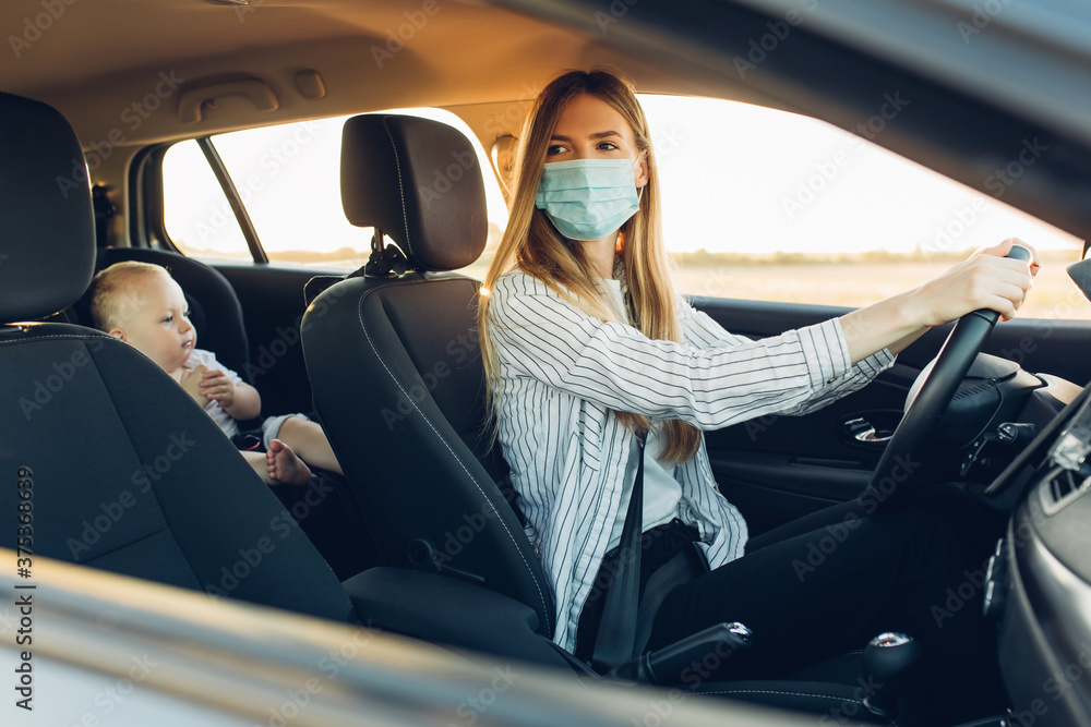 Young business woman in a protective medical mask on her face, driving a car during a trip with a small child in a car seat