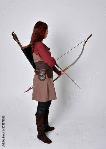 Full length portrait of girl with red hair wearing brown medieval archer costume.. Standing pose with back to the camera holding a bow and arrow, isolated against a grey studio background.