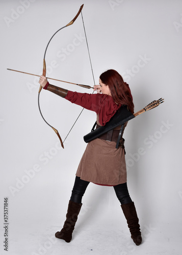 Full length portrait of girl with red hair wearing brown medieval archer costume.. Standing pose with back to the camera holding a bow and arrow, isolated against a grey studio background.