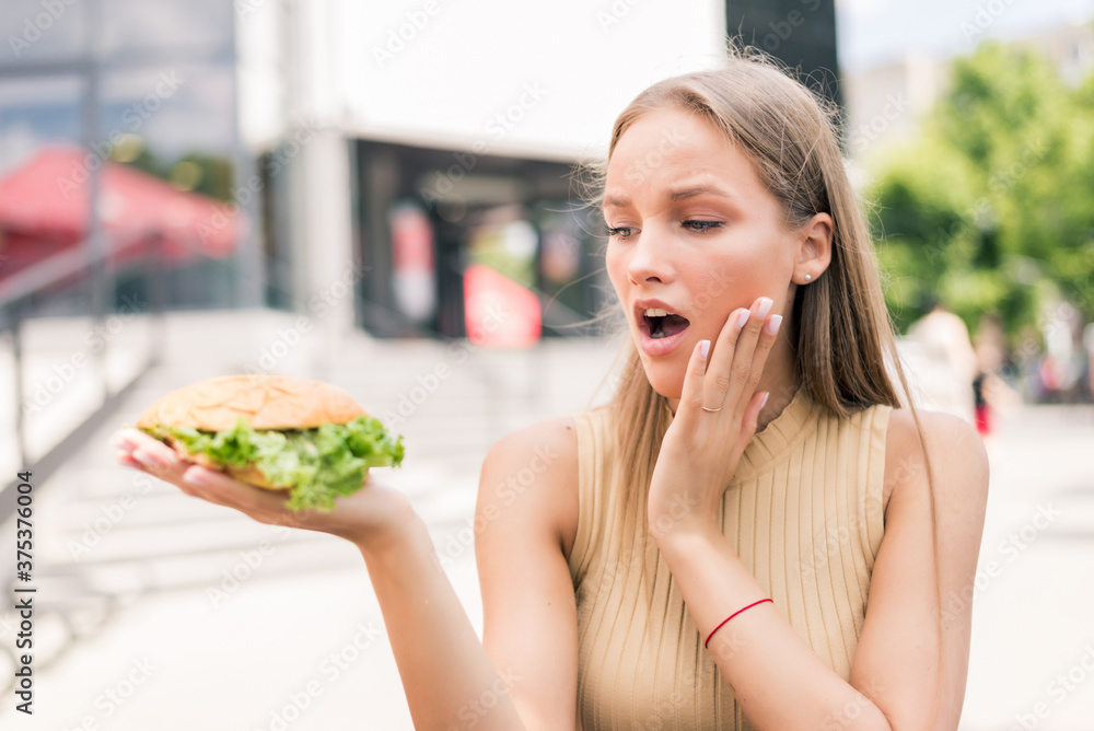 Young surprised woman holding burger while sitting in outdoors fast food Young surprised woman holding burger while sitting in outdoors fast food