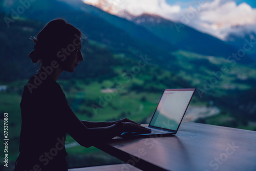 Skilled female digital nomad working remotely on freelance project sitting at table near big window with spectacular view of high mountains, young woman blogger messaging during networking on netbook © BullRun