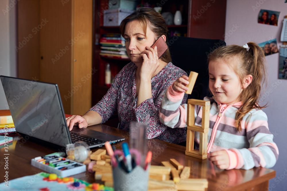 Fototapeta premium Woman mother working doing her job remotely during video chat call stream online course webinar on laptop from home while her daughter playing with bricks toy. Woman sitting at desk with computer