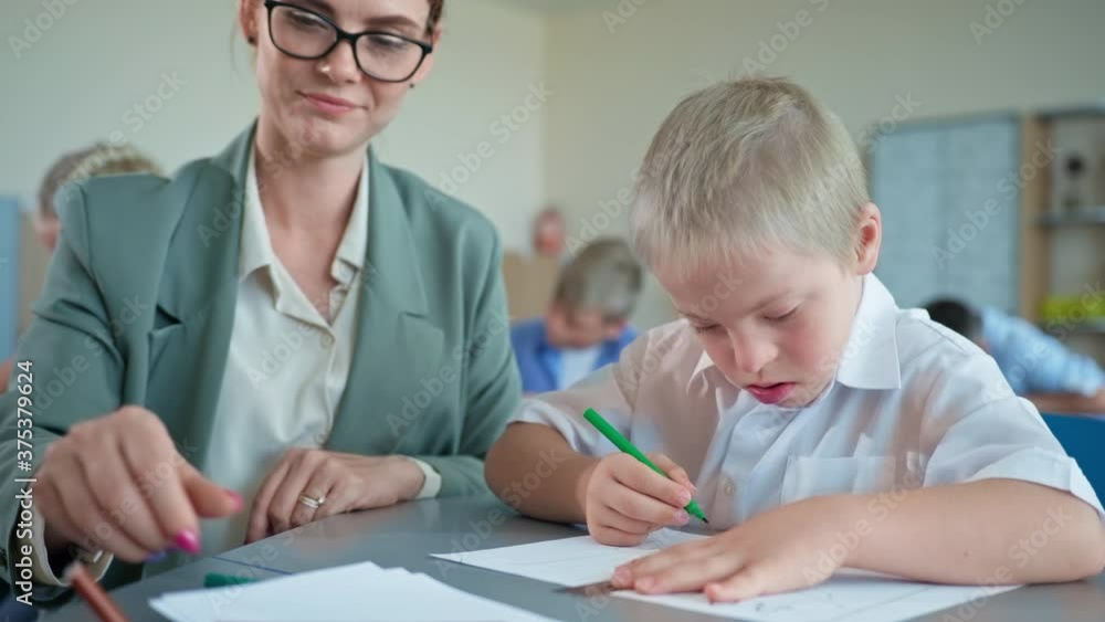 pedagogue works with a down syndrome boy, young woman in glasses teaches the pupil sitting at desk at grade school closeup pedagogue works with a down syndrome boy, young woman in glasses teaches the pupil sitting at desk at grade school closeup
