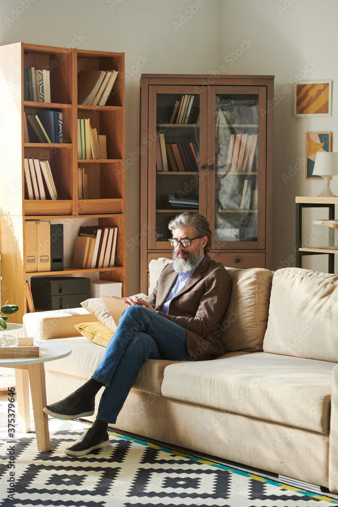Vertical shot of handsome bearded man in stylish outfit sitting relaxed on sofa in modern office room making notes
