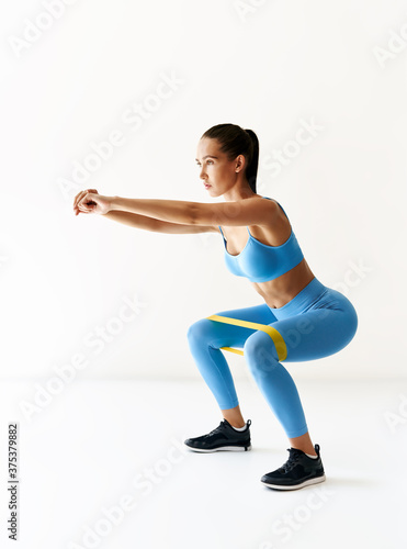 Sporty woman practicing squat exercises with resistance band loop over white background