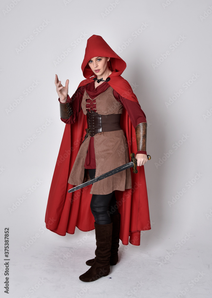 Obraz premium Full length portrait of girl wearing medieval costume and red cloak. Standing pose holding a sword, isolated against a grey studio background.