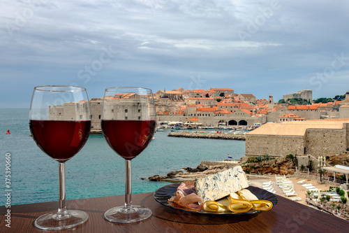 Fototapeta Naklejka Na Ścianę i Meble -  Two glasses of wine with charcuterie assortment against Dubrovnik city center background. Vacation concept. Beach and city wall in historic center of Dubrovnik, Croatia