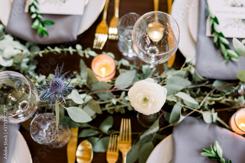 Top down view of a table with beautiful roses  eucalyptus leaves  and a greenery garland on a table set with empty wine glasses and gold silverware. 