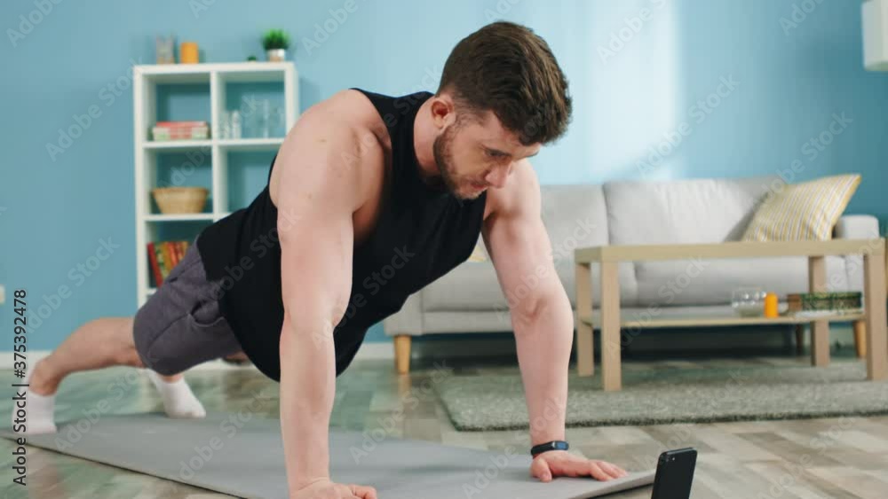 Young athletic man is standing in elbow plank, then changes to full arms plank, workouting in the morning at home, using stopwatch on a gadget, puts efforts to strengthen his body, Slow motion. Young athletic man is standing in elbow plank, then changes to full arms plank, workouting in the morning at home, using stopwatch on a gadget, puts efforts to strengthen his body, Slow motion.