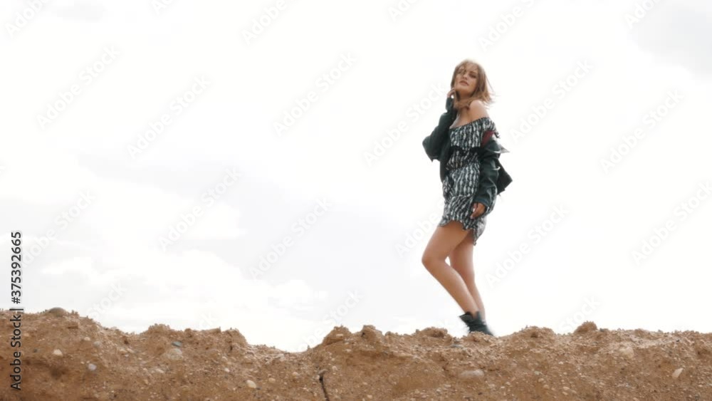 Happy attractive slim female model in dress and Leather Jacket walking on sandy mountain, woman brown hair blowing in wind. People and Nature Happy attractive slim female model in dress and Leather Jacket walking on sandy mountain, woman brown hair blowing in wind. People and Nature