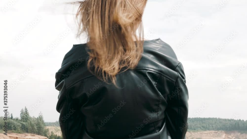 Back view of Young happy woman in Leather Jacket looking at sky and field, female brown hair blowing in wind. Feeling of freedom