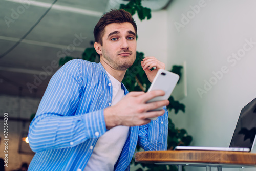 Portrait Of squeamish male freelancer looking at camera while using cellular and laptop for e learning in coworking space, confused Caucasian man with technology for doing distance job
