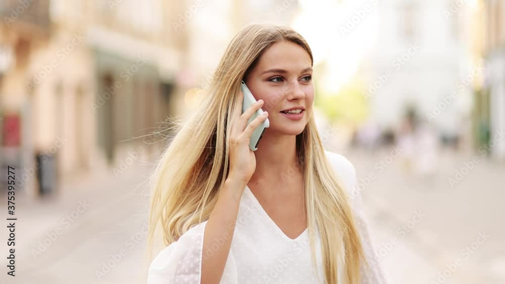 Slow motion front view of a young beautiful blonde woman in a white dress walking and calling on phone in the street. Slow motion front view of a young beautiful blonde woman in a white dress walking and calling on phone in the street.