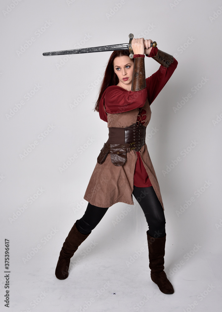 Full length portrait of girl wearing medieval costume. Standing pose holding a sword, isolated against a grey studio background. Full length portrait of girl wearing medieval costume. Standing pose holding a sword, isolated against a grey studio background.