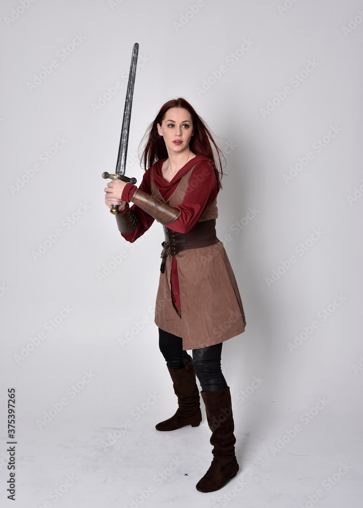 Full length portrait of girl wearing medieval costume. Standing pose holding a sword, isolated against a grey studio background. Full length portrait of girl wearing medieval costume. Standing pose holding a sword, isolated against a grey studio background.