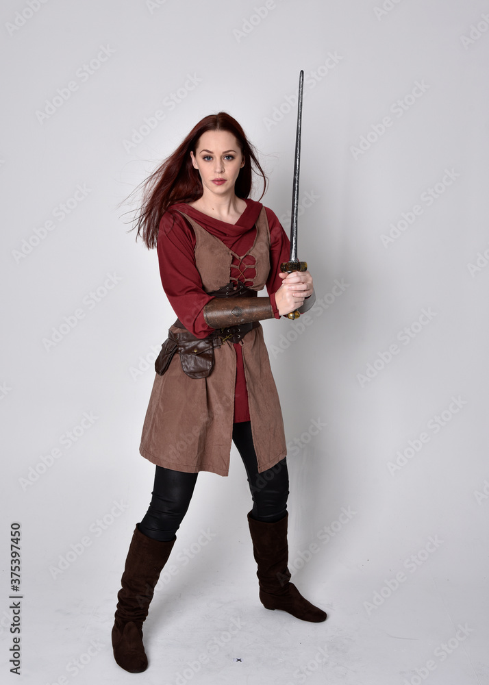 Full length portrait of girl wearing medieval costume. Standing pose holding a sword, isolated against a grey studio background. Full length portrait of girl wearing medieval costume. Standing pose holding a sword, isolated against a grey studio background.