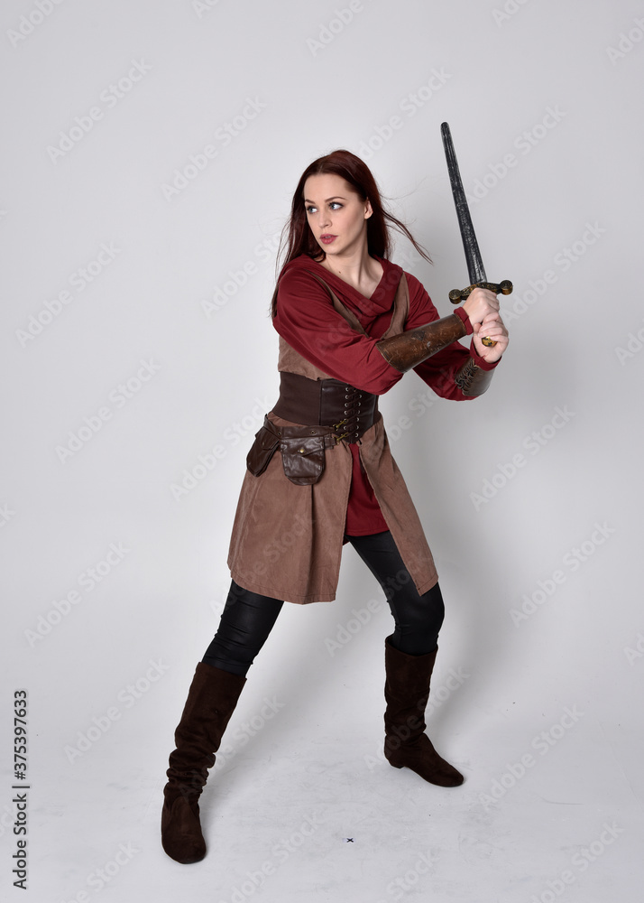 Full length portrait of girl wearing medieval costume. Standing pose holding a sword, isolated against a grey studio background. Full length portrait of girl wearing medieval costume. Standing pose holding a sword, isolated against a grey studio background.